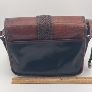 May include: A brown and black leather crossbody bag with a woven strap closure. The top flap has a crocodile texture, and the bottom is a glossy black. The bag has a silver-toned metal clasp and a brown leather strap.