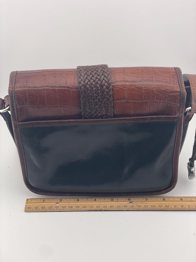 May include: A brown and black leather crossbody bag with a woven strap closure. The top flap has a crocodile texture, and the bottom is a glossy black. The bag has a silver-toned metal clasp and a brown leather strap.
