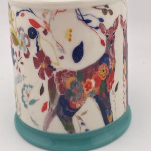 May include: A ceramic mug with a white background, featuring a deer design adorned with colorful floral patterns. The mug has a teal base and interior rim, with additional floral and leaf accents. The overall design is whimsical and artistic.