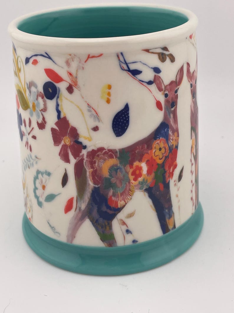 May include: A ceramic mug with a white background, featuring a deer design adorned with colorful floral patterns. The mug has a teal base and interior rim, with additional floral and leaf accents. The overall design is whimsical and artistic.