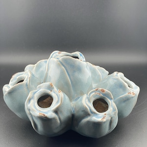 Puede incluir: Un jarrón de cerámica azul claro con forma de ramo de rosas. El jarrón tiene seis aberturas para flores, cada una con una forma ligeramente diferente.