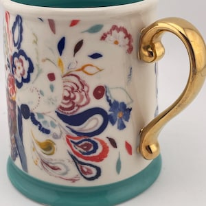 May include: A ceramic mug with a white base, adorned with colorful floral patterns in red, blue, and yellow. It features a gold-colored handle and a teal-colored rim and base. The mug is designed for beverages.