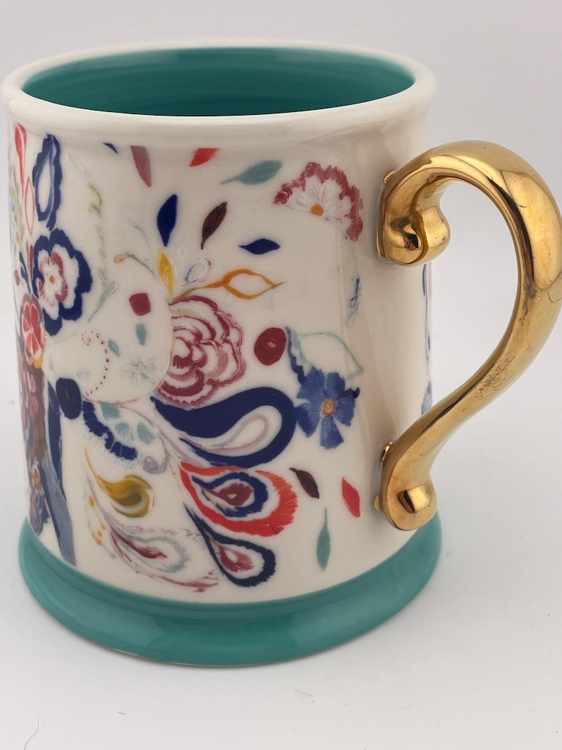 May include: A ceramic mug with a white base, adorned with colorful floral patterns in red, blue, and yellow. It features a gold-colored handle and a teal-colored rim and base. The mug is designed for beverages.