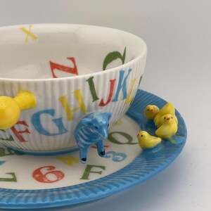 May include: A white ceramic teacup and saucer set with a blue rim. The teacup features colorful alphabet letters and a ridged design. The saucer has yellow chicks and a blue lamb figurine. A charming and whimsical design.