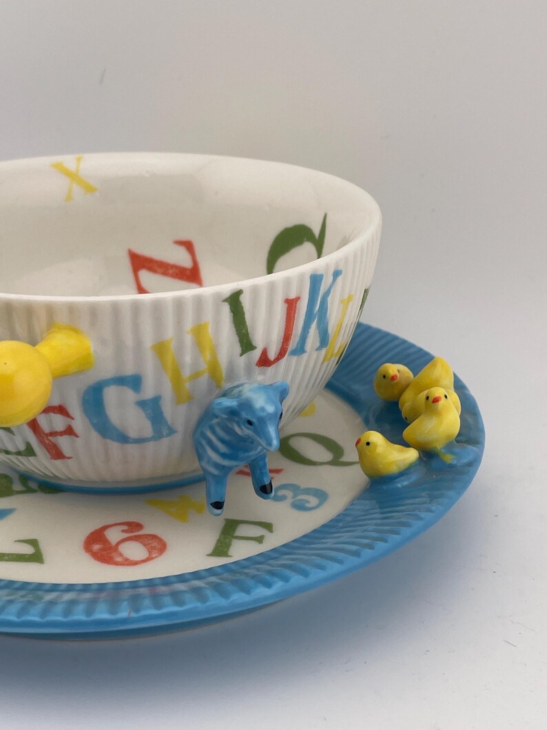May include: A white ceramic teacup and saucer set with a blue rim. The teacup features colorful alphabet letters and a ridged design. The saucer has yellow chicks and a blue lamb figurine. A charming and whimsical design.