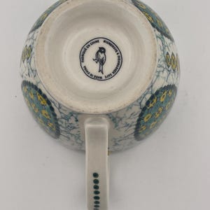 May include: White ceramic mug with a handle, featuring a blue and yellow floral pattern. The bottom of the mug has a circular stamp with text and a bird illustration. The handle is decorated with a series of small green dots.