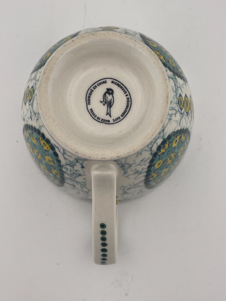 May include: White ceramic mug with a handle, featuring a blue and yellow floral pattern. The bottom of the mug has a circular stamp with text and a bird illustration. The handle is decorated with a series of small green dots.