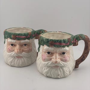 May include: Two ceramic mugs shaped like Santa Claus faces. Each mug features a sculpted white beard, pink cheeks, and a green wreath rim decorated with brown pinecones and red berries. The handles are textured to resemble brown tree branches.