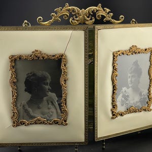 May include: An antique double picture frame with ornate gold-colored detailing. The frame holds two black and white portraits. The background is a cream color. The frame has a decorative top and stands on small feet.