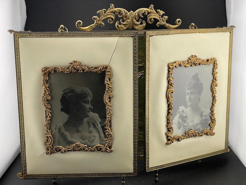 May include: An antique double picture frame with ornate gold-colored detailing. The frame holds two black and white portraits. The background is a cream color. The frame has a decorative top and stands on small feet.