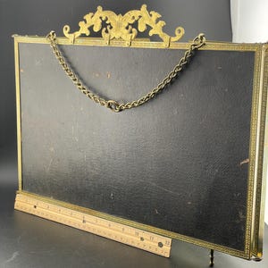 May include: An antique brass picture frame with a black backing. The frame has a decorative top and a chain for hanging. A wooden ruler is placed in front of the frame. The frame is approximately 12 inches long.