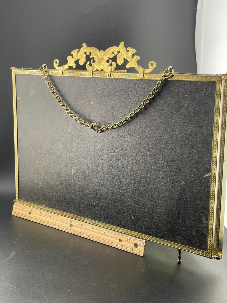 May include: An antique brass picture frame with a black backing. The frame has a decorative top and a chain for hanging. A wooden ruler is placed in front of the frame. The frame is approximately 12 inches long.
