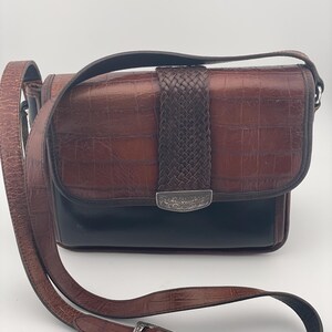 May include: A brown leather crossbody bag with a crocodile-effect texture. It has a woven leather detail and a silver-tone buckle on the adjustable strap. The bag's base is black. The strap is adjustable.