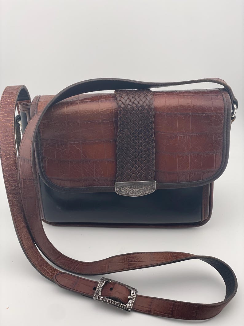 May include: A brown leather crossbody bag with a crocodile-effect texture. It has a woven leather detail and a silver-tone buckle on the adjustable strap. The bag's base is black. The strap is adjustable.