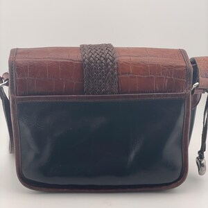 May include: A brown and black leather crossbody bag with a woven strap closure. The top section of the bag has a textured, crocodile-like pattern, while the bottom is smooth and glossy. It has a long, adjustable strap.