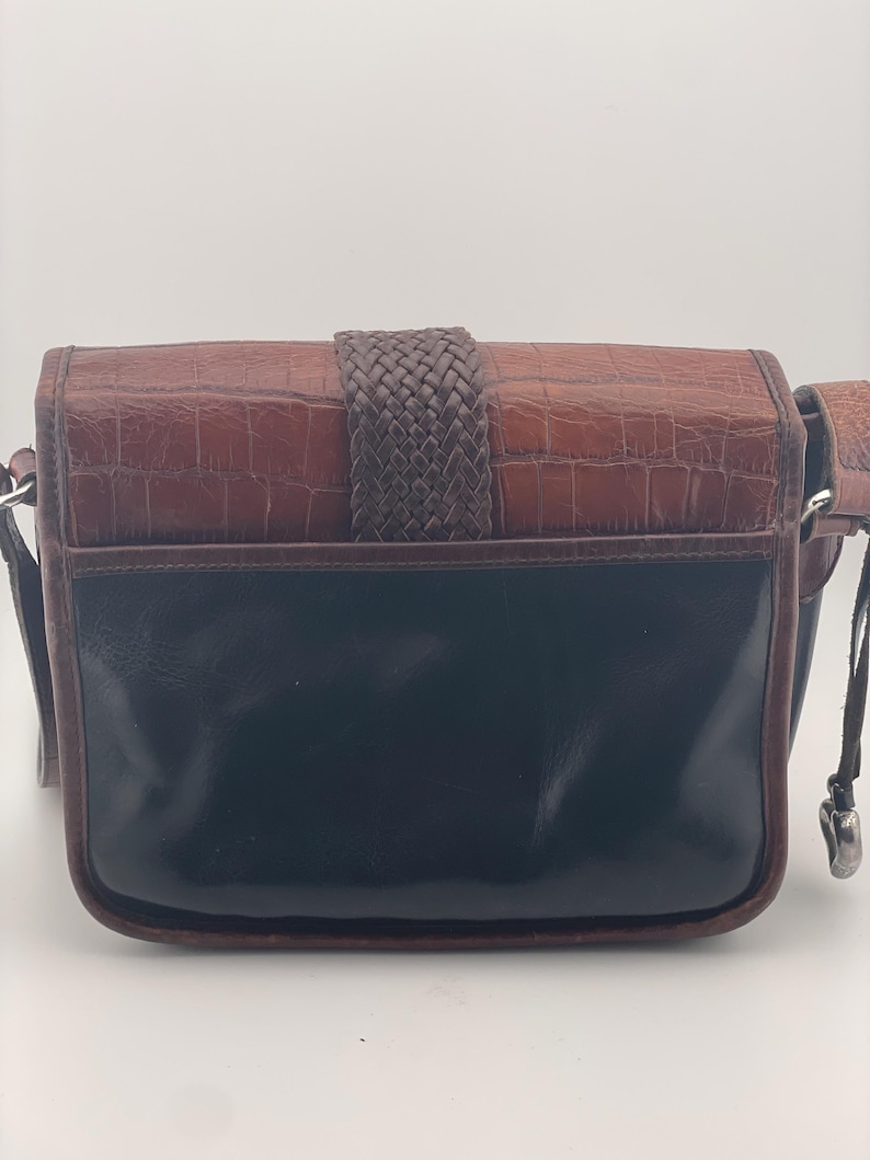 May include: A brown and black leather crossbody bag with a woven strap closure. The top section of the bag has a textured, crocodile-like pattern, while the bottom is smooth and glossy. It has a long, adjustable strap.