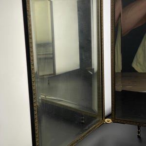 May include: A three-panel vanity mirror with a gold-colored decorative frame. The mirror is open, revealing reflections of the room. The frame has a detailed leaf pattern. The mirror is on a black surface.