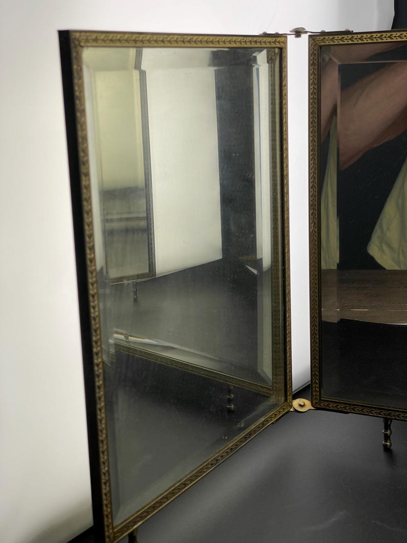 May include: A three-panel vanity mirror with a gold-colored decorative frame. The mirror is open, revealing reflections of the room. The frame has a detailed leaf pattern. The mirror is on a black surface.