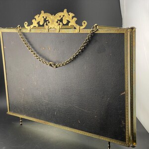 May include: An antique brass picture frame with a black backing. The frame has ornate detailing at the top and a chain for hanging. The frame is supported by small legs.