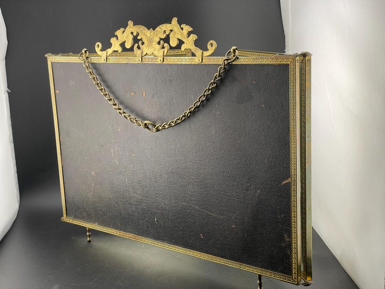May include: An antique brass picture frame with a black backing. The frame has ornate detailing at the top and a chain for hanging. The frame is supported by small legs.