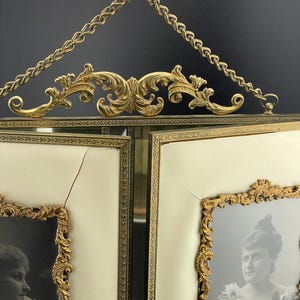May include: Antique double picture frame with ornate gold-tone filigree and chain. The frame holds two black and white portraits. The background is off-white.