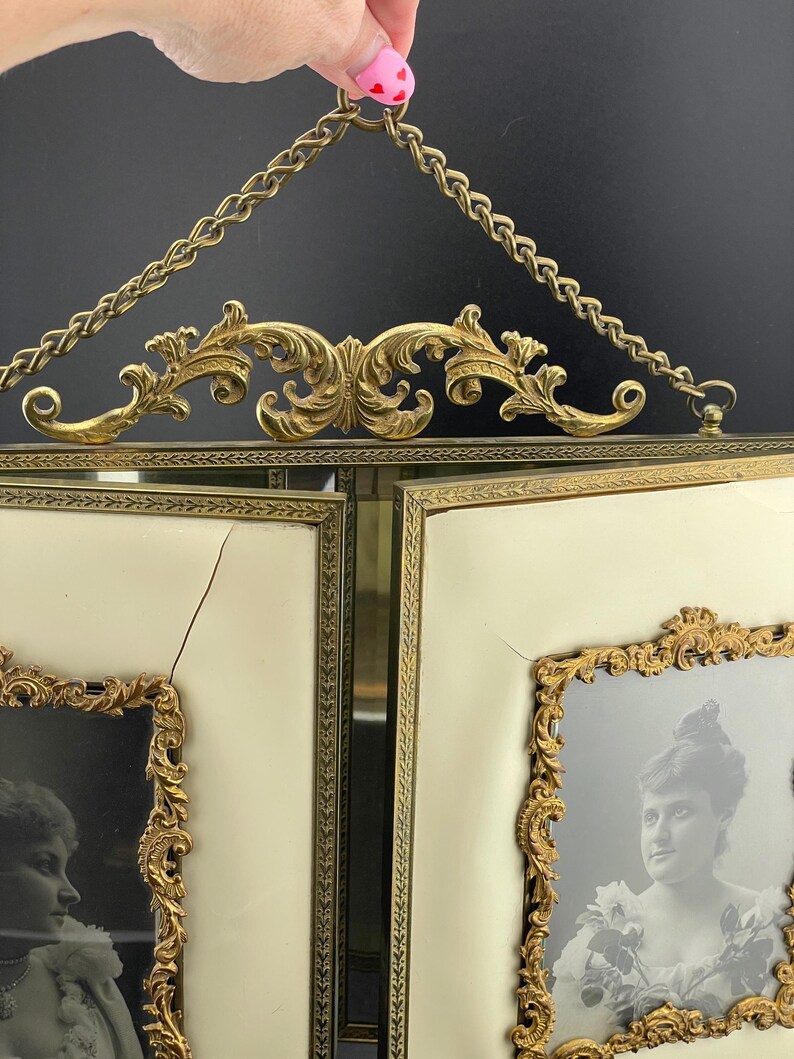 May include: Antique double picture frame with ornate gold-tone filigree and chain. The frame holds two black and white portraits. The background is off-white.
