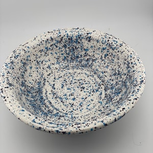 Puede incluir: Un cuenco de cerámica redondo con una base de color blanco y un patrón moteado de salpicaduras de pintura azul oscuro y negra en las superficies interior y exterior. El cuenco tiene una base ancha y poco profunda y un borde curvo.