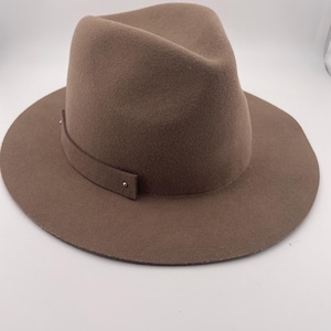 Puede incluir: Un sombrero fedora marr&oacute;n con ala ancha y una banda alrededor de la corona. El sombrero tiene una forma cl&aacute;sica y estructurada y est&aacute; hecho de un material suave similar al fieltro. La banda est&aacute; asegurada con un peque&ntilde;o detalle met&aacute;lico.