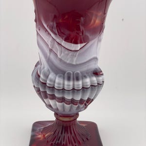 May include: A decorative glass vase with a square base and a flared top. The vase features a blend of red, white, and grey colours, with a ribbed design around the middle. The base is a deep red, and the overall style is vintage.