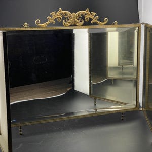 May include: A vintage vanity mirror with a gold-toned frame and ornate detailing. The mirror has three panels and a decorative top. The frame is accented with a gold filigree design. The mirror is supported by small, decorative legs.