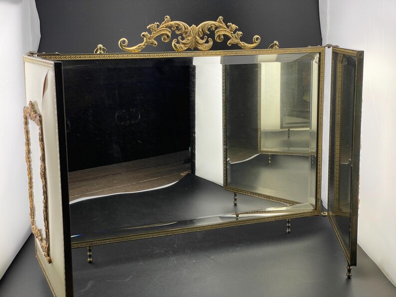 May include: A vintage vanity mirror with a gold-toned frame and ornate detailing. The mirror has three panels and a decorative top. The frame is accented with a gold filigree design. The mirror is supported by small, decorative legs.