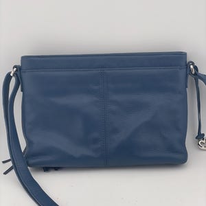 May include: A blue leather crossbody bag with a long adjustable strap and silver-toned hardware. The bag has a rectangular shape with a front pocket and a top zipper closure. The bag's strap is 48 inches long.