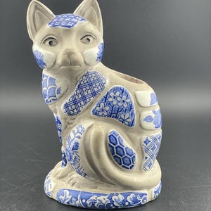 Puede incluir: Una figurita de cerámica de un gato con un diseño de patrón azul y blanco. El gato está sentado con la cabeza girada hacia un lado y la cola enroscada alrededor de su cuerpo.