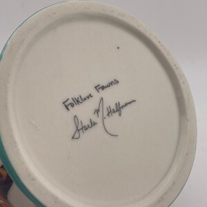 May include: A ceramic vase with a white base and a turquoise rim. The bottom of the vase features the handwritten text "Folklore Fawns" and a signature. The vase has a colorful design on the side.