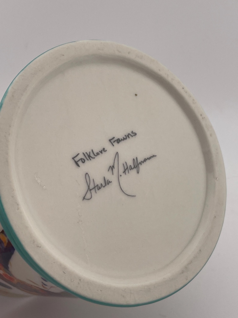 May include: A ceramic vase with a white base and a turquoise rim. The bottom of the vase features the handwritten text "Folklore Fawns" and a signature. The vase has a colorful design on the side.