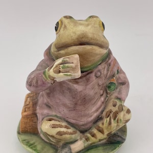 Op de afbeelding: Een keramieken beeldje van een kikker in een paarse jas, met een klein vierkant voorwerp. De kikker zit op een groene, bladvormige basis. Het beeldje heeft een grillige, antieke esthetiek.
