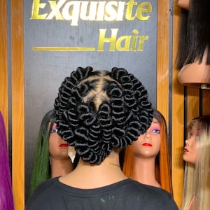 May include: A person with black hair styled in a heart shape with tight curls. The person is wearing a black shirt and is standing in front of a wall with mannequins with different colored wigs.
