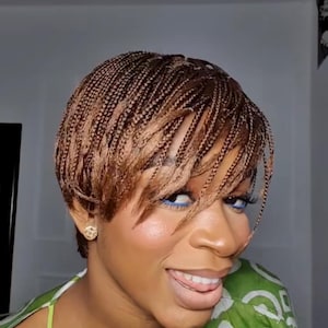 Peut inclure: Une femme avec une coiffure courte tressée dans des tons bruns et blonds. Les tresses sont étroitement tissées, créant un aspect texturé. Elle porte des boucles d'oreilles dorées et a un eye-liner bleu. La femme sourit et porte un haut vert et blanc.