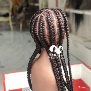 Peut inclure: Une perruque brune avec un style tressé, comportant cinq rangées de tresses avec un total de six tresses. La perruque est sur une tête de mannequin et les tresses sont longues et épaisses. La perruque est étiquetée "SARAH EXQUISITE HAIR".