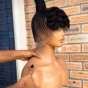 Puede incluir: Una peluca negra trenzada, peinada en una cabeza de maniquí. La peluca presenta una trenza alta y cilíndrica en la coronilla y un diseño de trenzas decorativas en capas. El maniquí está frente a una pared de ladrillos.