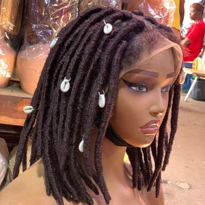 May include: A dark brown wig featuring shoulder-length faux locs styled with white cowrie shell accents. The wig has a lace front construction and is displayed on a mannequin head, showcasing the textured, cylindrical strands of the hair extensions.