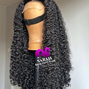 May include: A black curly wig on a mannequin head. The wig has a lace front and a black elastic band. The hair is long and voluminous, with tight curls. The text "SARAH EXQUISITE HAIR" is visible on the stand.