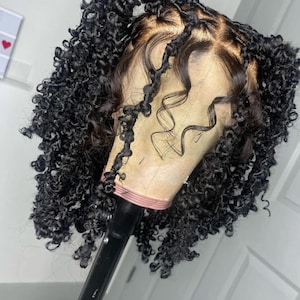 May include: A black curly wig styled on a mannequin head. The wig features a lace front and a natural-looking hairline. The curls are tightly coiled and voluminous, creating a full and textured appearance. The wig is mounted on a black stand.