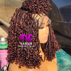 May include: A brown braided wig with curly ends and a bun on top. The wig is on a mannequin head. The text "SARAH EXQUISITE HAIR" is visible. A can of hairspray is in the background.