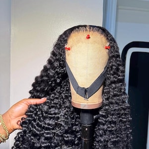 May include: A black curly wig styled on a mannequin head. The wig features a natural hairline and is secured with red pins. The hair is a deep black color and has a voluminous, curly texture. The mannequin head is mounted on a black stand.