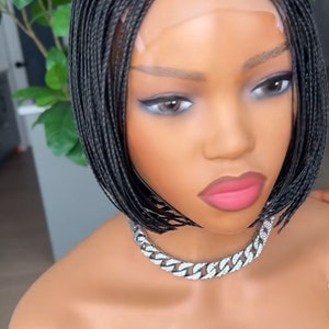 Op de afbeelding: Een zwarte gevlochten pruik in bob-stijl. De pruik zit op een mannequinhoofd met een zilveren kettingketting. De tekst "Sarahexquisitehair" is zichtbaar onderaan de afbeelding.