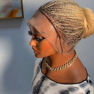 May include: A blonde braided wig styled in a bun, worn on a mannequin head. The wig is secured with a lace front. A gold chain necklace is worn around the neck. The mannequin is wearing a black and white tie-dye top.