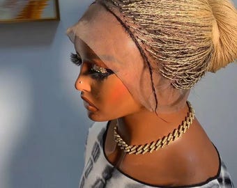 May include: A blonde braided wig styled in a bun, worn on a mannequin head. The wig is secured with a lace front. A gold chain necklace is worn around the neck. The mannequin is wearing a black and white tie-dye top.