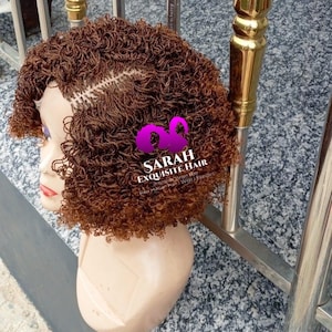Peut inclure: Perruque bouclée marron sur une tête de mannequin. La perruque présente une coupe courte et texturée avec des racines plus foncées et des pointes plus claires. Le texte "SARAH EXQUISITE HAIR" est visible.