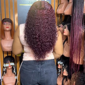 May include: A woman wearing a burgundy curly wig in front of a wall of mannequin heads with various hairstyles. The sign above the heads reads "Sarah Exquisite".
